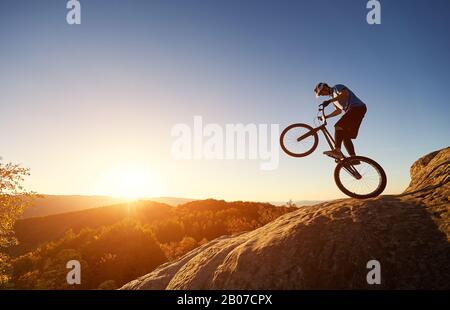 Silhouette of cyclist balancing on back wheel on trial bicycle. Courageous sportsman biker making acrobatic trick on top of rocky mountain at sunset. Concept of extreme sport active lifestyle Stock Photo