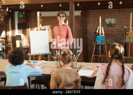 Young female teacher teaching children to paint while they sitting at the table during art lesson in art studio Stock Photo