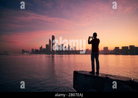 Young man with mobile phone photographing urban skyline at sunrise. Abu Dhabi, United Arab Emirates. Stock Photo