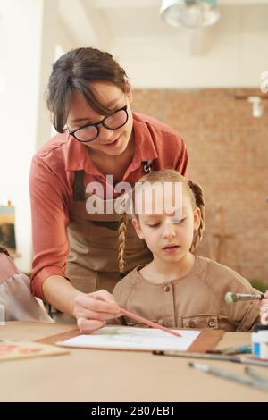 Young woman holding paintbrush and helping little girl to paint during a lesson Stock Photo