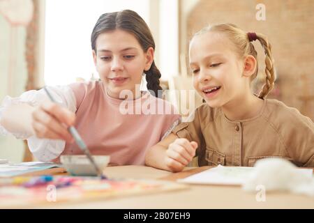 Two happy friends sitting at the table and painting a picture together with paints at school Stock Photo