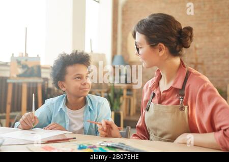 Young woman in eyeglasses talking to African little boy while they sitting at the table and painting during art lesson at school Stock Photo