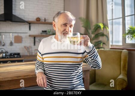 Bearded grey-haired man in a striped sweater having tea Stock Photo