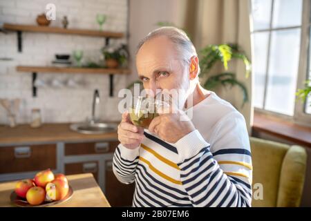 Bearded grey-haired man in a striped sweater having tea in the kitchen Stock Photo