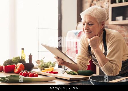 Elderly woman using digital tablet in kitchen, checking cooking recipes Stock Photo