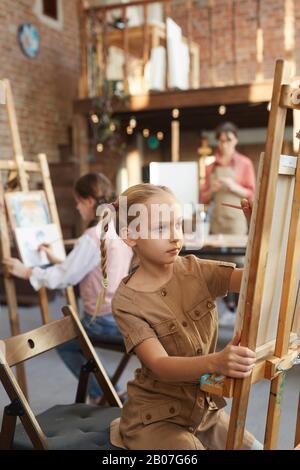 Little girl sitting in front of easel and painting with other students during art lesson at art studio Stock Photo