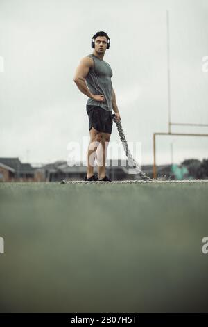 Strong young man standing in the field with a battle rope and looking away. Sportsman taking break from battling rope workout outdoors. Stock Photo