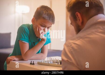 young man and son playing chess together. Son is thinging about his next move. Stock Photo