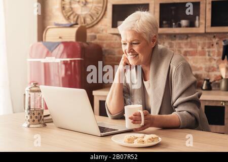 Cheerful senior woman using laptop in kitchen and drinking coffee Stock Photo