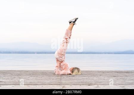A beautiful woman performs the exercises lifting her legs to the top, lake, pier, Fitness. Sport. Yoga. Stock Photo
