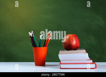 Back to school, orange pencil holder, stack of books on white table with red apple, empty green school board background, education concept Stock Photo