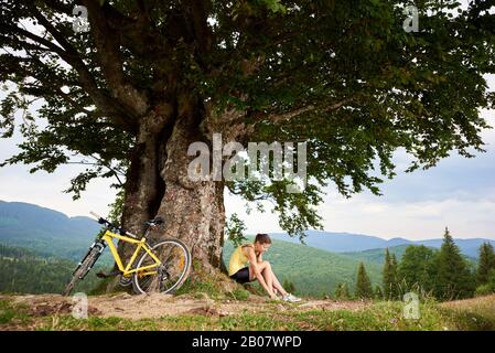 Attractive happy woman cyclist sitting near yellow mountain bicycle under big tree, enjoying summer day in the mountains. Outdoor sport activity, lifestyle concept Stock Photo