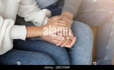 Unrecognizable Couple Holding Hands Sitting On Couch Indoor, Cropped Stock Photo