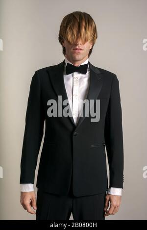 Young man with long shaggy hair standing in formal tuxedo with shirt and black tie Stock Photo