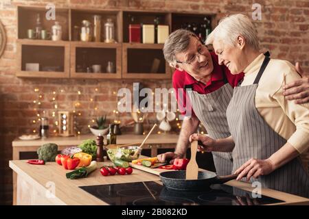 Happy Aged Couple Embracing And Cooking Dinner Together In Kitchen Stock Photo