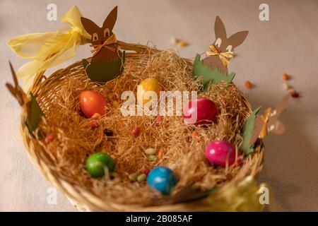 Paper rabbit are sitting around colorful cooked easter eggs and candy eggs in hay basket. Close up, high angle view. Stock Photo