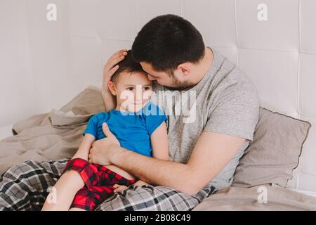 Young Caucasian father talking to boy son. Man parent hugging with child on bed in bedroom at home. Authentic lifestyle real candid moment. Happy Fath Stock Photo