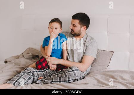 Young Caucasian father talking to boy son. Man parent hugging with child on bed in bedroom at home. Authentic lifestyle real candid moment. Happy Fath Stock Photo
