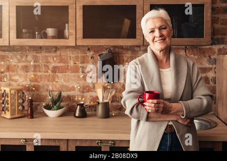 Happy elderly woman standing in kitchen with cup of coffee Stock Photo