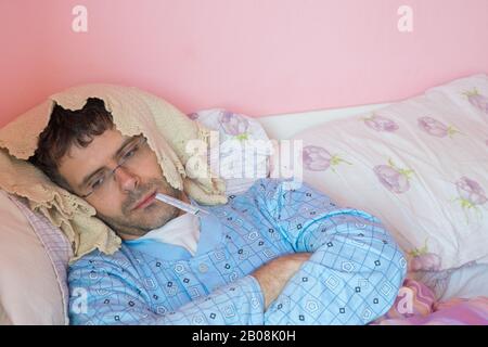 Portrait of a sick young man lying in bed taking temperature and having flu. Stock Photo