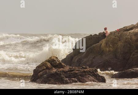 A fisherman sits with a wooden fishing pole on a rocky promontory hit by the high tidal waves of waters of the sea in Gokarna. Stock Photo