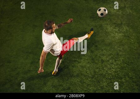 Top view of caucasian football or soccer player on green background of grass. Young male sportive model training, practicing. Kicking ball, attacking, catching. Concept of sport, competition, winning. Stock Photo