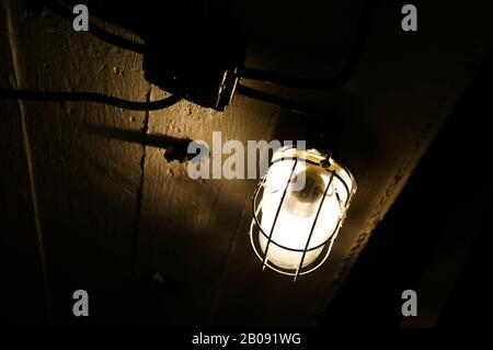 Close up of old isolated damp-proof luminaire with grid shining from dark cellar ceiling Stock Photo
