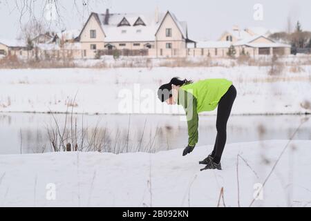 Young brunette woman in green jacket standing at lake and doing exercise before running Stock Photo