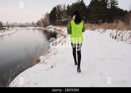 Rear view of woman in green jacket running along winter shore with long dry grass Stock Photo