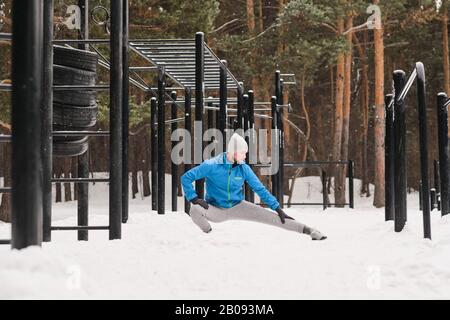 Young man in warm clothes doing crouch adductor stretch while exercising at winter sports ground Stock Photo