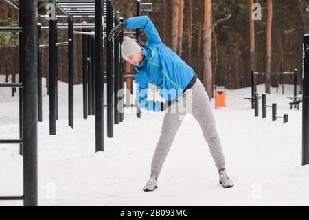 Sporty young man in hat standing at workout area in winter and doing warm-up exercise Stock Photo