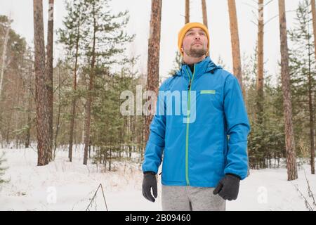 Handsome young man in blue jacket looking around and enjoying winter stroll in forest Stock Photo
