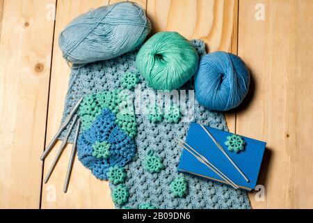 Composition of knitting tools and crocheted blue-green openwork napkins, balls of colored yarn, plastic grey hooks and book on wooden background. Conc Stock Photo