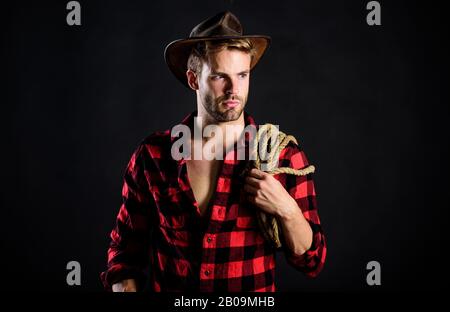 Lassoing on prairie. Man unshaven cowboy black background. Cowboy wearing hat hold rope. Lasso tool of American cowboy. Still used on ranches to capture cattle or other livestock. Western life. Stock Photo