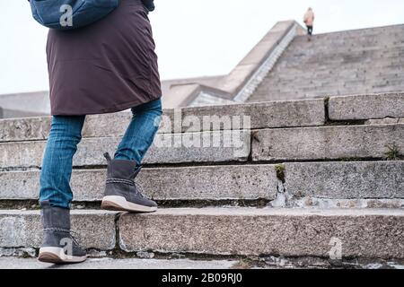 Tourist in warm clothes and with an urban backpack walks up the old stone steps of a high staircase on a winter day. Stock Photo