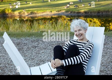 beautiful, happy Senior woman sitting on Adirondack chair in park with pond and geese Stock Photo