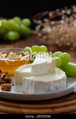 White, round cheeses on wooden background with honey, nuts and grapes. Dark food photo. Stock Photo