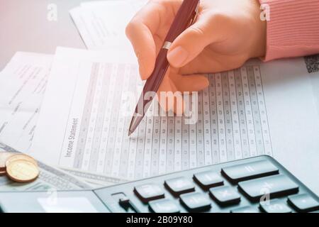 Close Up side view on the hand of accountant girl who is holding a pen and looking in a document with a numerical table for the necessary data. Profit Stock Photo