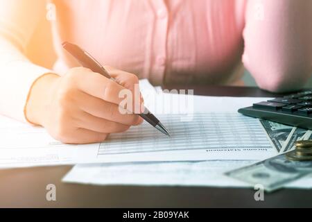 Closeup front view on the hand of a girl who is holding a pen and doing calculations using documents with tables and a calculator. Calculation of taxe Stock Photo