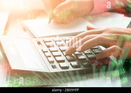 Female accountant works in the office using a calculator and writes the results in a notebook. Double exposure with digital graphics and a workplace. Stock Photo