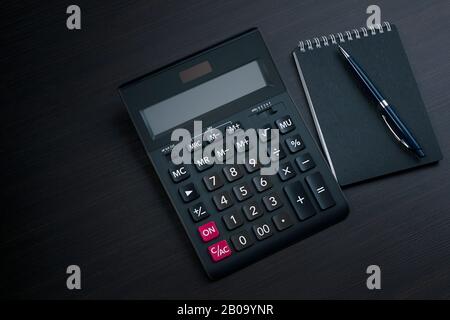 Top view on black calculator with blank screen, pen and notebook on a dark wooden table with copy space. Profit and market analysis concept Stock Photo