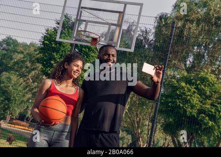 Outdoors Activity. African couple standing on basketball court boyfriend taking selfie on smartphone with girlfriend smiling toothy Stock Photo