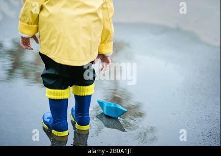 Little boy in raincoat and rubber boots playing in puddle. Happy little kid with paper ship by a puddle on warm summer day after rain. Active leisure Stock Photo