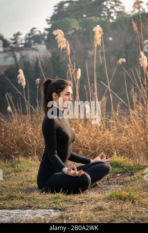 Portrait of woman doing yoga on the bank of a river with shrubs Stock Photo