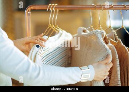 Closeup on trendy 40 years old woman in white sweater and skirt in the modern house in sunny winter day choosing sweaters hanging on copper clothes ra Stock Photo