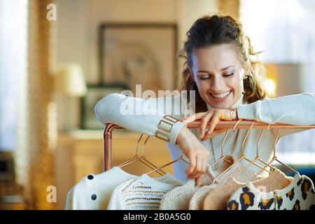 cheerful young woman in white sweater and skirt at modern home in sunny winter day choosing sweaters hanging on copper clothes rack. Stock Photo