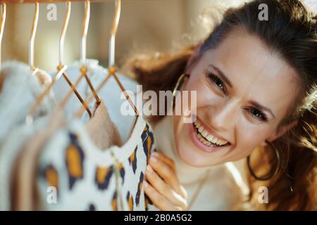 Portrait of happy young woman in white sweater and skirt in the modern house in sunny winter day choosing sweaters hanging on copper clothes rack. Stock Photo