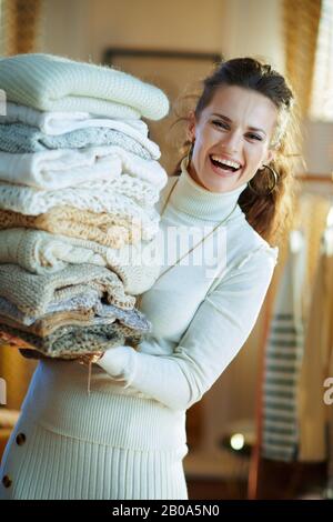 cheerful stylish 40 years old housewife in white sweater and skirt in the modern house in sunny winter day holding huge pile of sweaters. Stock Photo