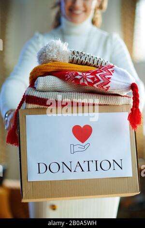 Closeup on smiling modern female in white sweater and skirt giving donation box with old warm clothes in the modern living room in sunny winter day. Stock Photo