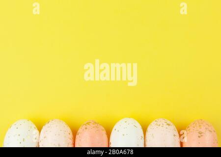 Creatively painted eggs in pastel colors decorated with gold leaf are arranged in row along bottom edge on yellow background, copy space. Happy Easter Stock Photo